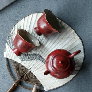 May include: A tea set consisting of a round, dark red teapot with a lid and two matching tea cups with a textured, dark red exterior and a smooth, light-coloured interior. The teapot and cups are arranged on a decorative, circular paper fan.