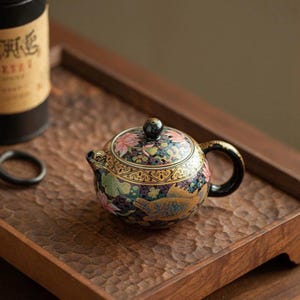 Peut inclure: Une petite théière ronde en céramique à base noire, ornée de motifs peints à la main représentant des poissons dorés, des fleurs de lotus roses et des nénuphars verts. Elle possède un couvercle assorti avec un bouton et une anse noire courbée.