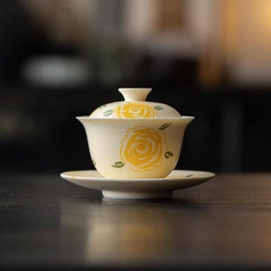 May include: A cream-coloured ceramic gaiwan tea set consisting of a bowl, a lid, and a saucer. The set features a hand-painted yellow rose design with small green leaves on the exterior of the bowl and the top of the lid, with a smooth finish.