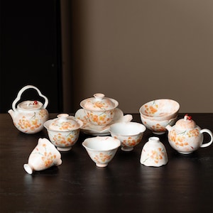 May include: A ceramic tea set featuring a white base with hand-painted orange and yellow floral patterns. The set includes two teapots with arched handles, lidded bowls, and several small cups, all decorated with delicate blossoms and red accents.