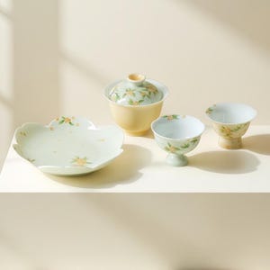 Puede incluir: Un juego de té de cerámica que consta de un cuenco con tapa, dos tazas con pie y un plato festoneado. Cada pieza presenta una base de color amarillo pálido y blanco con un delicado patrón de pequeñas flores naranjas y hojas verdes.