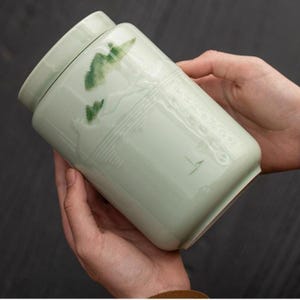 May include: A cylindrical ceramic tea canister with a pale celadon green glaze. The exterior features a subtle embossed landscape pattern with hand-painted green accents depicting mountain peaks and a small bird, topped with a matching lid.