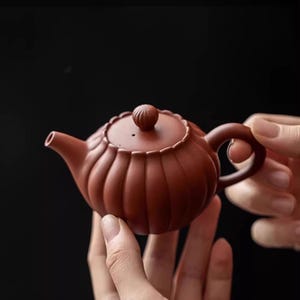 Könnte beinhalten: Eine rotbraune Tonteekanne mit einem geriffelten, kürbisartigen Körper und einem passenden Deckel mit einem kleinen, gerippten Knopf. Die Teekanne hat einen geschwungenen Griff und einen kurzen, konischen Ausguss mit matter Oberfläche.