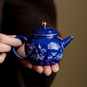 Op de afbeelding: Een kleine, ronde theepot van donkerblauw keramiek met een wit geschilderd patroon van delicate vlinders en bloemranken. De theepot heeft een bijpassend deksel met een kleine, glanzende goudkleurige knop aan de bovenkant en een tuit.