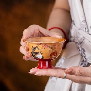 Puede incluir: Un pequeño cuenco de té de cerámica con borde acampanado y base de pedestal roja sólida. El exterior presenta un esmalte de color marrón dorado cálido decorado con una ilustración pintada a mano de dos gatos y flores de cerezo rosadas.