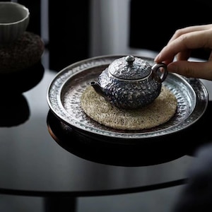 May include: A small, dark, metallic teapot with a textured, hammered surface and a matching lid. The teapot rests on a round, fibrous, tan-colored mat, which is placed inside a wide, circular, silver-toned metal tray with a decorative rim.