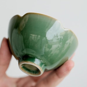 Puede incluir: Un pequeño cuenco de cerámica festoneado con un esmalte brillante de color verde salvia intenso. El cuenco presenta una superficie lisa y reflectante con un pie circular distintivo en la base. El borde tiene una forma ondulada y sutil.