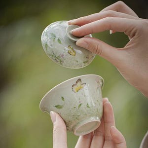 May include: A light green ceramic tea bowl with a matching lid, featuring a delicate hand-painted pattern of yellow butterflies, green leaves, and small pink flowers. The bowl has a smooth, rounded shape with a flared rim and a small knob on the lid.
