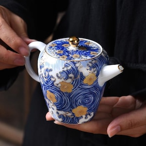 May include: A white ceramic teapot featuring a blue swirling wave pattern and gold floral accents. The teapot has a matching lid with a gold spherical knob, a curved white handle, and a short spout with a gold-trimmed rim.