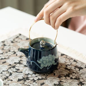 Puede incluir: Una tetera de cerámica azul oscuro con un patrón floral azul claro en el cuerpo, un asa de metal arqueada en tono dorado y un pomo de tapa decorativo en tono plateado. La tetera tiene un acabado liso y brillante y un pico corto.