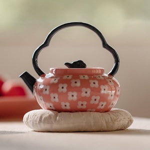 May include: A ceramic teapot with a coral pink base color featuring a repeating pattern of small white flowers with dark centers. The teapot has a black spout, a black lid handle, and a tall, arched black handle with a distinct curved shape.