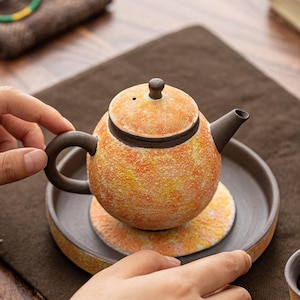 Puede incluir: Una tetera de cerámica redonda con un acabado texturizado y moteado en naranja y amarillo, con asa, pico y tapa de color marrón oscuro. La tetera descansa sobre un posavasos circular a juego, colocado dentro de una bandeja circular marrón oscuro.