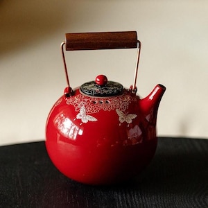 Könnte beinhalten: Eine runde, glänzend rote Keramik-Teekanne mit einem weißen Schmetterlings- und Blumenmuster. Sie verfügt über einen schwarzen Deckel mit rotem Knauf, einen kurzen Ausguss und einen Metallgriff mit einem dunkelbraunen Holzgriffstück.