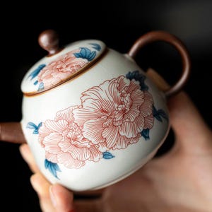 May include: A round ceramic teapot with a cream-colored base featuring a fine crackle glaze. The surface is decorated with detailed red line-art peony flowers and small blue leaves. It includes a matching lid with a brown knob and a brown handle.