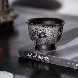 May include: A black ceramic tea cup featuring a detailed silver-toned illustration of a fox surrounded by swirling patterns and small pink cherry blossom accents. The cup has a flared rim and a pedestal base, showcasing a textured matte finish.