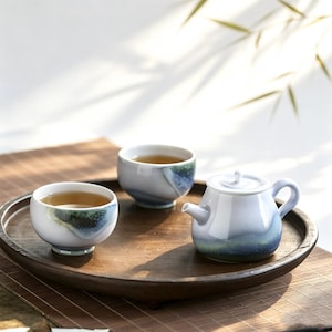 May include: A ceramic tea set consisting of a small teapot and two matching cups, all featuring a smooth, light blue glaze with dark blue and green mottled accents at the base. The pieces are arranged on a round, dark brown wooden serving tray.