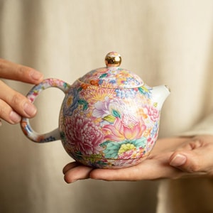 以下が含まれることがあります： ピンク、黄色、青、緑の花々が描かれた鮮やかなマルチカラーのフローラルパターンが特徴の丸い陶器製ティーポット。ティーポットには、金色のつまみが付いたお揃いの蓋、湾曲したハンドル、短い注ぎ口があり、全体に花が装飾されています。