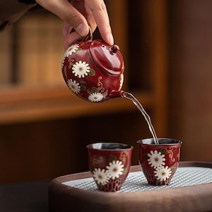 Può includere: Una teiera in ceramica rosso scuro con coperchio abbinato e due piccole tazze, tutte decorate con fiori bianchi simili a margherite, foglie verdi e viticci dorati. La teiera versa un liquido trasparente in una delle tazze su un vassoio di legno.