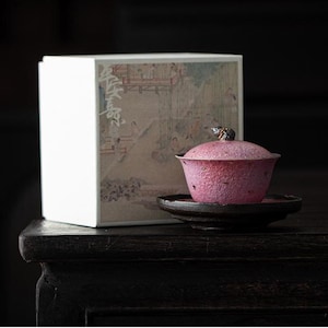 May include: A pink textured tea bowl with a matching lid and a small dark finial, resting on a dark circular saucer. Beside it sits a white rectangular box featuring a printed illustration of figures in a traditional architectural setting.