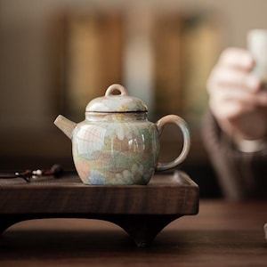 May include: A ceramic teapot with a glossy, crackled glaze featuring a soft, multi-coloured pastel pattern of pink, blue, and green hues. The teapot has a rounded body, a curved handle, a short spout, and a matching lid with a small loop handle.
