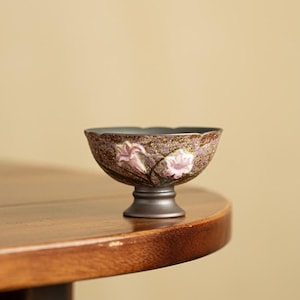 May include: A small, footed ceramic bowl featuring a textured, speckled brown and purple exterior. The bowl is decorated with two raised, light pink floral motifs on the side and has a smooth, dark grey pedestal base and a scalloped rim.