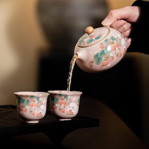 Puede incluir: Una tetera de cerámica rosa claro y dos tazas a juego decoradas con fresas rojas pintadas, hojas verdes y conejos blancos. La tetera tiene un pomo de madera redondo en la tapa y vierte té en una de las tazas, que presentan bordes festoneados.