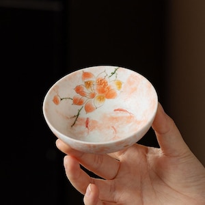 May include: A small, round ceramic tea bowl with a white base and a hand-painted orange lotus flower design. The interior features delicate orange brushstrokes and small fish motifs, creating a soft, watercolor-like appearance on the glazed surface.