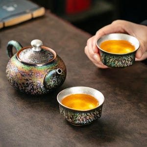 Peut inclure: Une théière en céramique et deux tasses assorties présentant un vernis irisé sombre avec des taches multicolores. La théière a un corps arrondi, un bec verseur incurvé et un couvercle argenté. Chaque tasse contient un thé jaune doré éclatant.