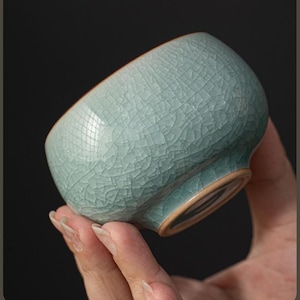 Puede incluir: Una pequeña taza de té de cerámica redondeada con un esmalte verde celadón pálido y un patrón de grietas intrincado y prominente en la superficie exterior. La base y el borde interior revelan una textura de arcilla marrón claro sin esmaltar.