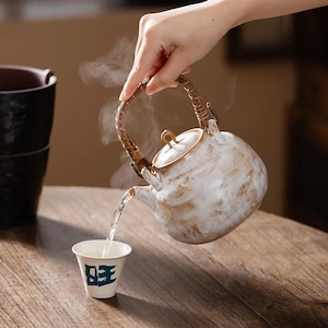 Puede incluir: Una tetera de cerámica con esmalte jaspeado blanco y marrón y un asa de bambú trenzado, vertiendo líquido caliente en una pequeña taza de cerámica blanca con un carácter chino azul oscuro en el frente, con vapor visible saliendo del pico.
