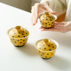 Puede incluir: Tres cuencos de té de cerámica pequeños con tapas a juego, cada uno con un color base amarillo vibrante decorado con un patrón de lunares negros oscuros. Los cuencos tienen una forma redondeada con un borde acampanado y un pomo pequeño.