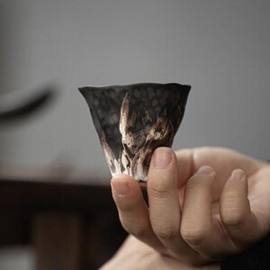 May include: A small, tapered ceramic tea cup featuring a dark, textured exterior with a mottled black and charcoal finish. The surface displays irregular, organic white and brown streaks that resemble natural stone patterns, with a flared rim.