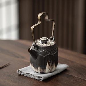 May include: A dark grey ceramic teapot with a textured, petal-like surface and white brushstroke accents at the base. It features a tall, arched metallic handle and a matching lid with a small, textured knob, showcasing a distinct artisan aesthetic.