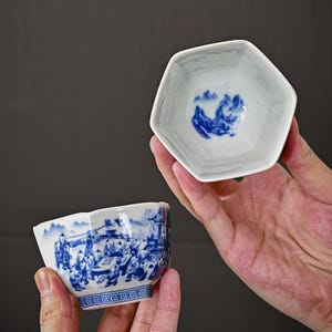 May include: A hexagonal ceramic tea cup featuring a white base with intricate blue underglaze illustrations of children playing in a landscape on the exterior and a mountain scene painted on the interior base, finished with a geometric border.