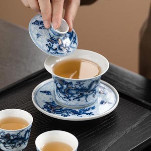 May include: A white porcelain gaiwan tea set featuring intricate blue dragon motifs and cloud patterns. The set includes a lidded bowl, a matching saucer, and two small tea cups, all filled with a light amber tea liquid.