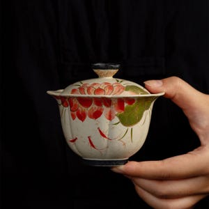 Peut inclure: Un bol à thé gaiwan en céramique avec couvercle assorti, présentant une base couleur crème ornée de fleurs de lotus rouges, de feuilles vertes et de petits poissons rouges peints à la main. Le bol a un bord cannelé et une base sombre.