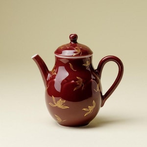 Op de afbeelding: Een diepbordeauxrode keramische theepot met een glanzende afwerking en een herhalend patroon van delicate goudkleurige florale lijntekeningen. De theepot is voorzien van een bijpassend deksel met een knop, een gebogen tuit en een ronde handgreep.