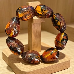 May include: A circular bracelet composed of eight polished, irregular amber-colored beads featuring deep brown, black, and golden-yellow marbled inclusions. The beads are strung together in a continuous loop, displaying a smooth, glossy finish.