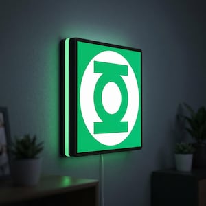 May include: A square wall-mounted LED sign featuring the Green Lantern emblem, which consists of a white circle containing a stylised green lantern symbol on a solid green background, with a black frame and a glowing green light border.