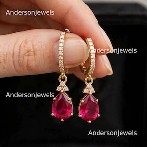 Puede incluir: Un par de pendientes de aro dorados con una hilera de pequeñas piedras claras a lo largo del aro y un colgante de piedra roja en forma de pera acentuado con tres pequeñas piedras claras. El texto AndersonJewels es visible.