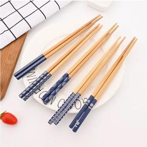 Peut inclure: Cinq paires de baguettes en bois brun clair disposées sur une assiette blanche. Chaque paire présente un manche bleu foncé distinct orné de motifs géométriques, floraux ou circulaires blancs. Les pointes sont effilées et texturées.