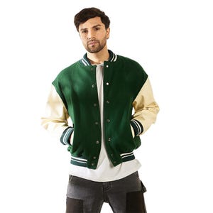 Puede incluir: Una chaqueta tipo varsity con cuerpo de lana en color verde oscuro y mangas color crema. La chaqueta cuenta con cierre frontal de botones a presión, cuello, puños y dobladillo acanalados con rayas blancas y verdes, y dos bolsillos laterales.