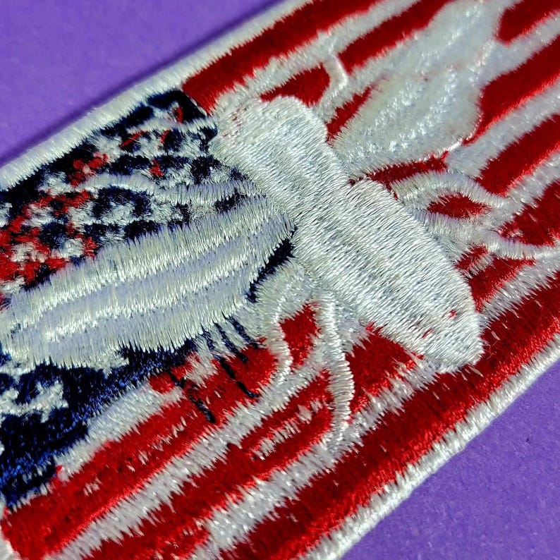 May include: A rectangular embroidered patch featuring a stylized white moth centered over a background of red and white horizontal stripes and a blue field with white stars, mimicking the pattern of the American flag with stitched texture.