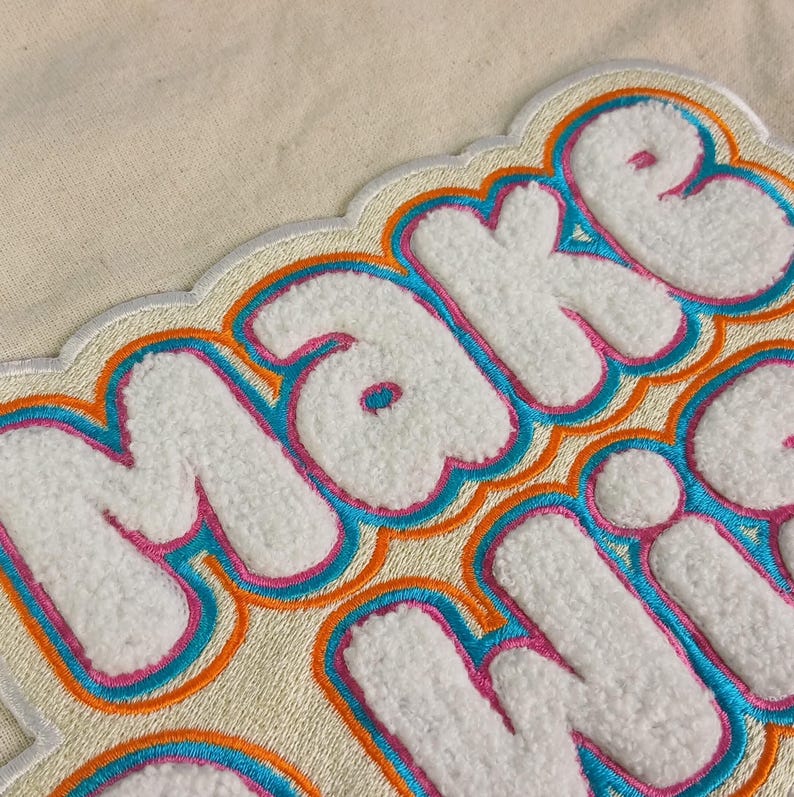 Op de afbeelding: Een witte stoffen patch met de tekst MAKE WISH in verhoogde, pluizige witte chenille letters. Elke letter is omlijnd met lagen felroze, levendig blauw en oranje borduurgaren, wat zorgt voor een opvallend, meerkleurig randeffect op de letters.