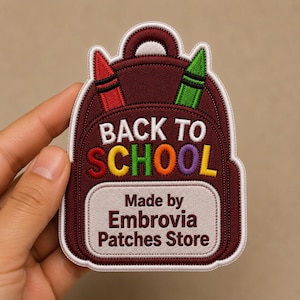 Puede incluir: Un parche bordado de color granate con forma de mochila, con un lápiz rojo y uno verde en la parte superior. El texto dice BACK TO SCHOOL en letras blancas y multicolores, con Made by Embrovia Patches Store en granate sobre fondo claro.