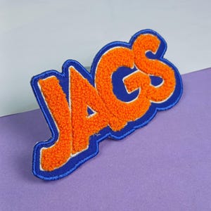 Op de afbeelding: Een geborduurde patch met het woord JAGS in feloranje, getextureerde letters in chenille-stijl. De letters zijn omlijnd met een dikke, gladde, koningsblauwe rand, wat zorgt voor een gedurfde, verhoogde uitstraling voor op textiel.