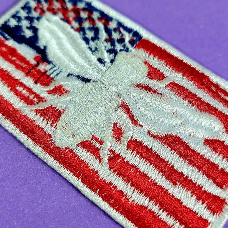 May include: A rectangular embroidered patch featuring a white bee silhouette centered over a background of red and white horizontal stripes and a blue field with white stars, mimicking the design of the American flag in a stitched textile format.