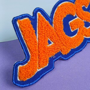 Op de afbeelding: Een geborduurde patch met de tekst JAGS in feloranje chenille letters. De letters zijn omlijnd met wit en geplaatst op een donkerblauwe stoffen achtergrond met een bijpassende blauwe gestikte rand, wat zorgt voor een getextureerd uiterlijk.