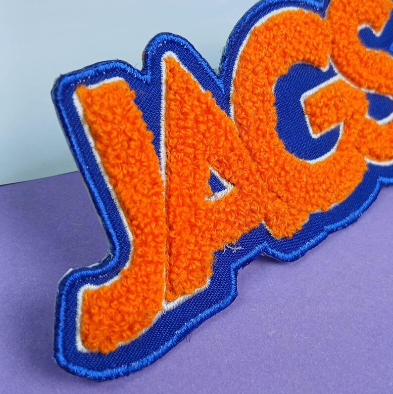 Op de afbeelding: Een geborduurde patch met de tekst JAGS in feloranje chenille letters. De letters zijn omlijnd met wit en geplaatst op een donkerblauwe stoffen achtergrond met een bijpassende blauwe gestikte rand, wat zorgt voor een getextureerd uiterlijk.
