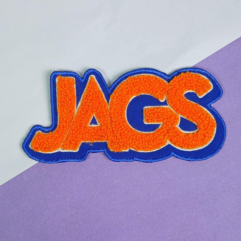Op de afbeelding: Een geborduurde patch met de tekst JAGS in feloranje chenille letters met een dikke, getextureerde pool. De letters zijn omlijnd met een gedurfde, gladde koningsblauwe rand die de contouren van het woord volgt, wat een gelaagd effect cre&euml;ert.
